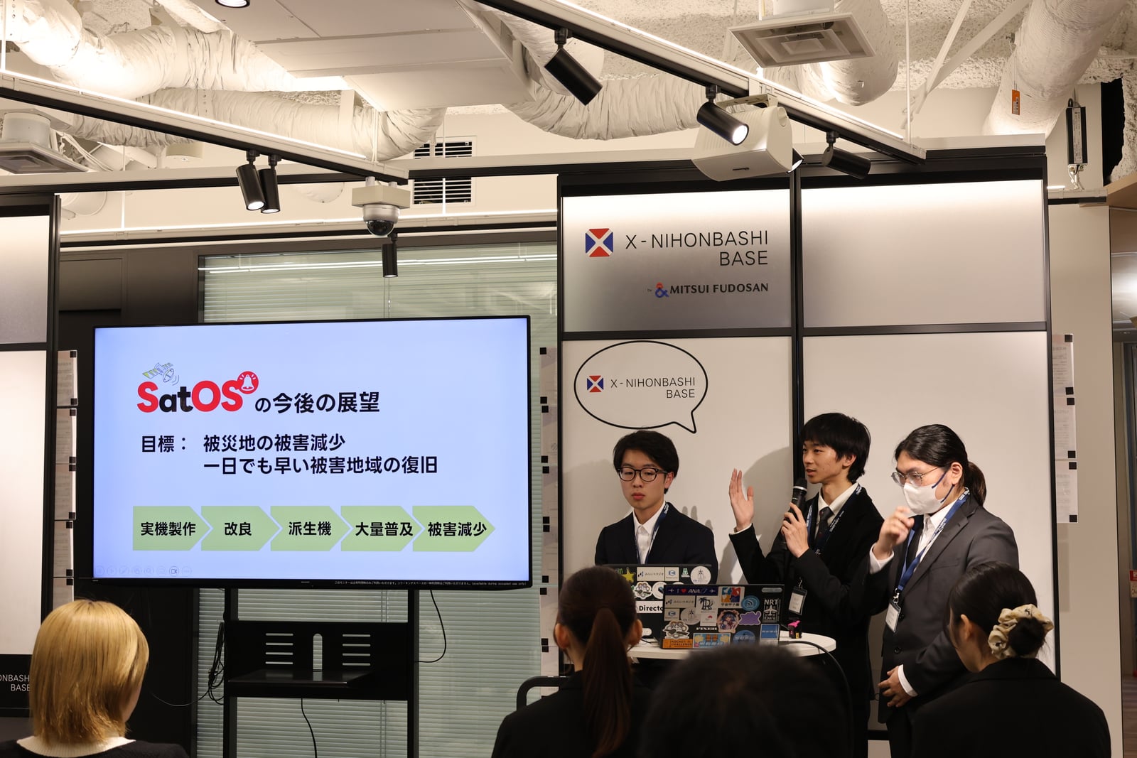 沖縄高専チームによる SatOS の最終発表。スライドには「SatOS の今後の展望：被災地の被害減少／一日でも早い被害地域の復旧」が映る